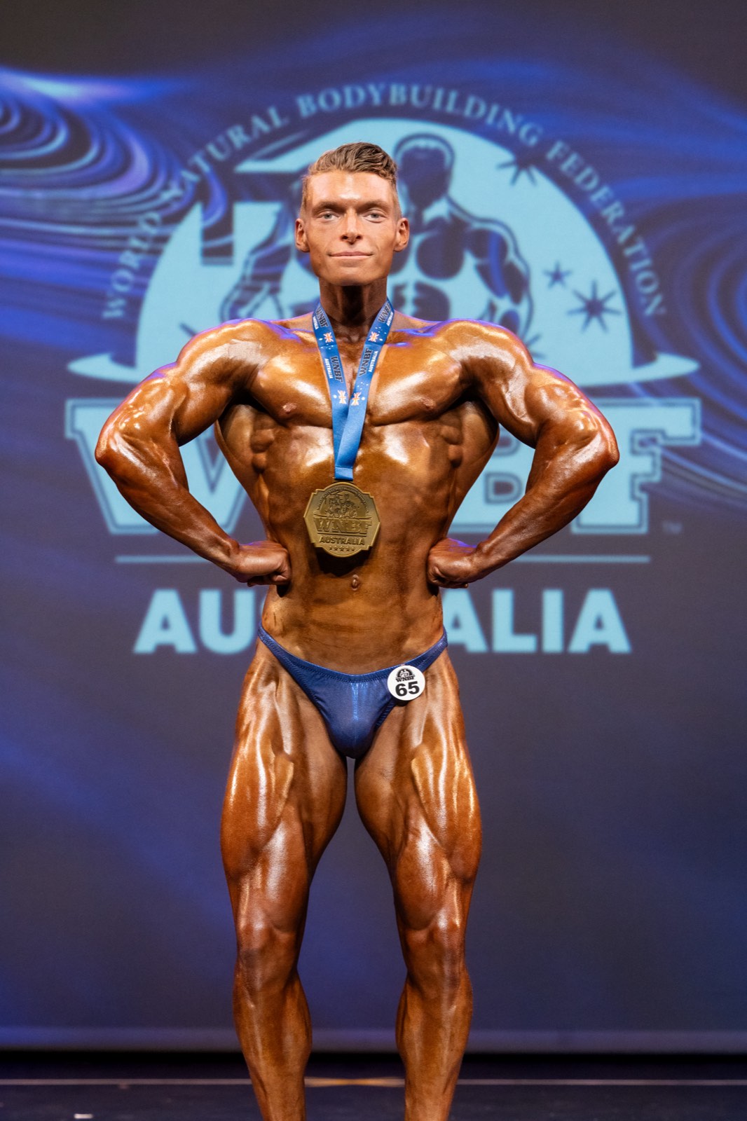 Bodybuilder number 65 in a front lat spread on stage, wearing his WNBF Australia gold medal