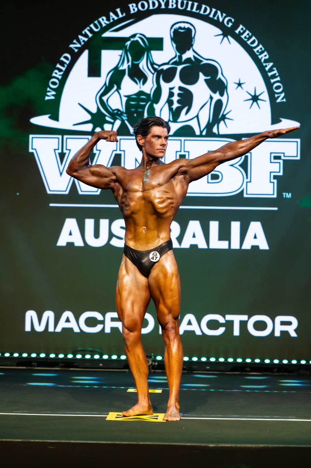 Athlete number 47 flexing one arm and extending the other in a pose on stage in front of the WNBF Australia backdrop