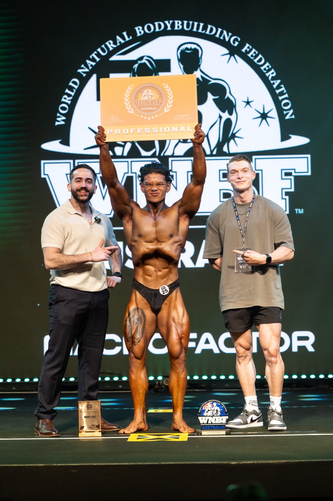 Classic physique athlete number 25 holding a WNBF Professional card above his head, with a presenter on each side pointing at him